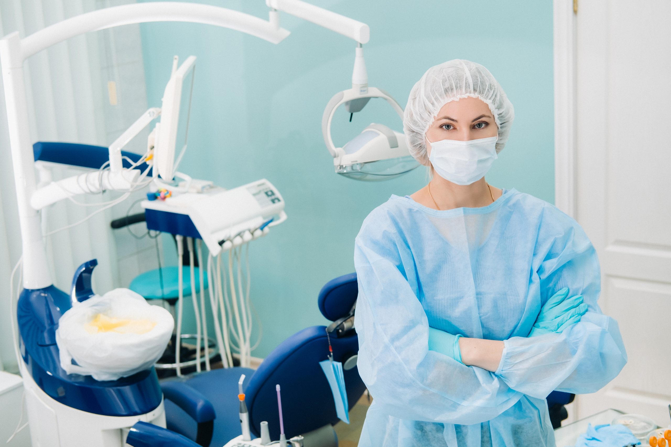 a female dentist wearing a medical mask and rubber gloves poses for the camera and folds her arms in her office